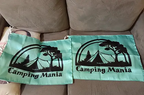 Dua bendera custom Camping Mania berwarna hijau muda dengan ilustrasi tenda dan pepohonan berwarna hitam, hasil cetak tajam dan finishing rapi.
