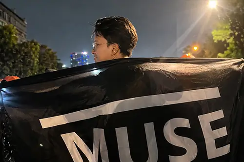 Seorang pelanggan membentangkan bendera custom berwarna hitam dengan tulisan MUSE pada acara malam hari, menampilkan hasil cetak yang tajam dan berkualitas.