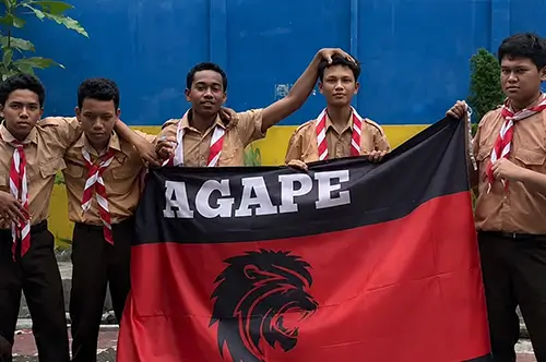 Sekelompok anggota Pramuka memegang bendera custom AGAPE berwarna hitam dan merah dengan logo kepala singa, hasil cetak tajam dan warna yang kontras.