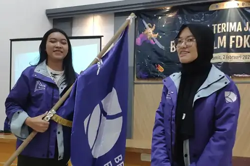 Dua mahasiswa memegang bendera custom berwarna ungu saat kegiatan kampus di dalam ruangan, dengan hasil cetak warna tajam dan finishing rapi dari Nusaflag.